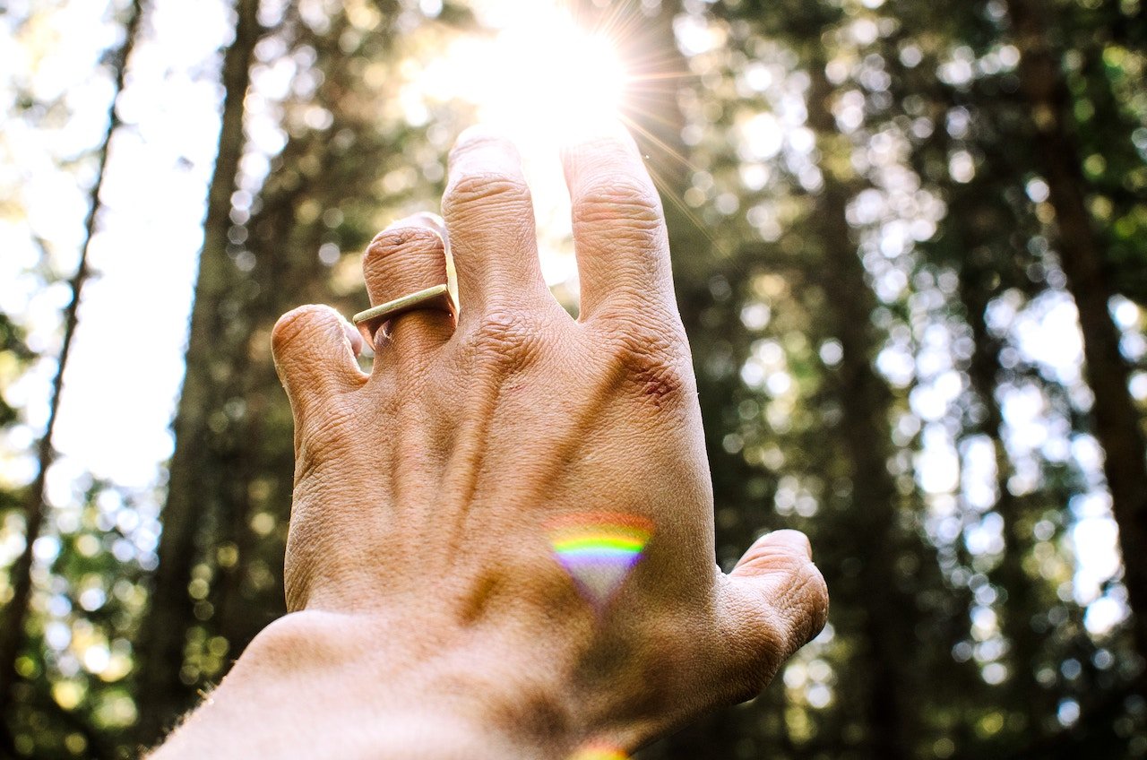 hand reaching for the sun shining through trees, with small rainbow reflection